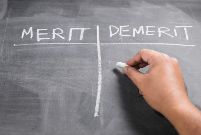 黒板に「MERIT（メリット）」と「DEMERIT（デメリット）」の比較表がチョークで書かれており、人の手がチョークを持って記入しようとしている様子。外国人雇用における採用と定着のメリット・デメリットを整理するイメージに最適。