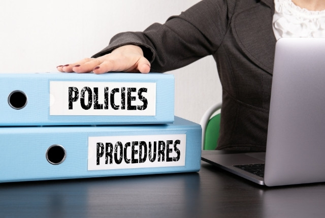 デスクに置かれた“Policies”“Procedures”とラベルの付いた青いファイルとノートパソコン、書類に手を添える人物