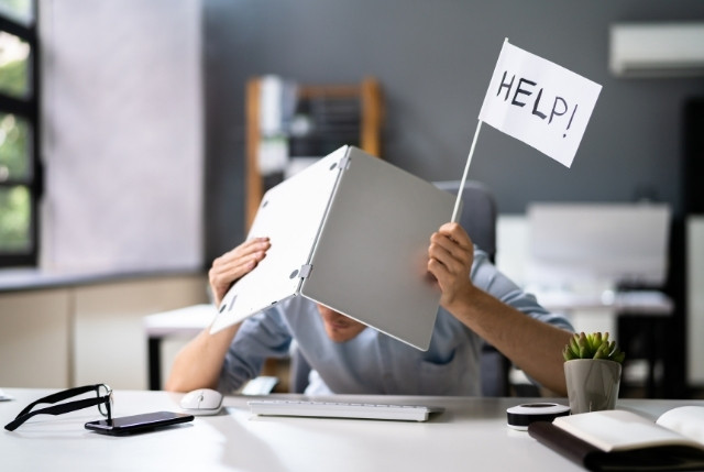オフィスでノートパソコンを頭にかぶり、手に『HELP!』と書かれた旗を持つ人物。仕事の困難や助けを求める状況を表すイメージ